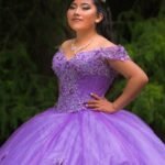 A woman elegantly posing in a detailed purple gown with a tiara, exhibiting glamour and confidence.